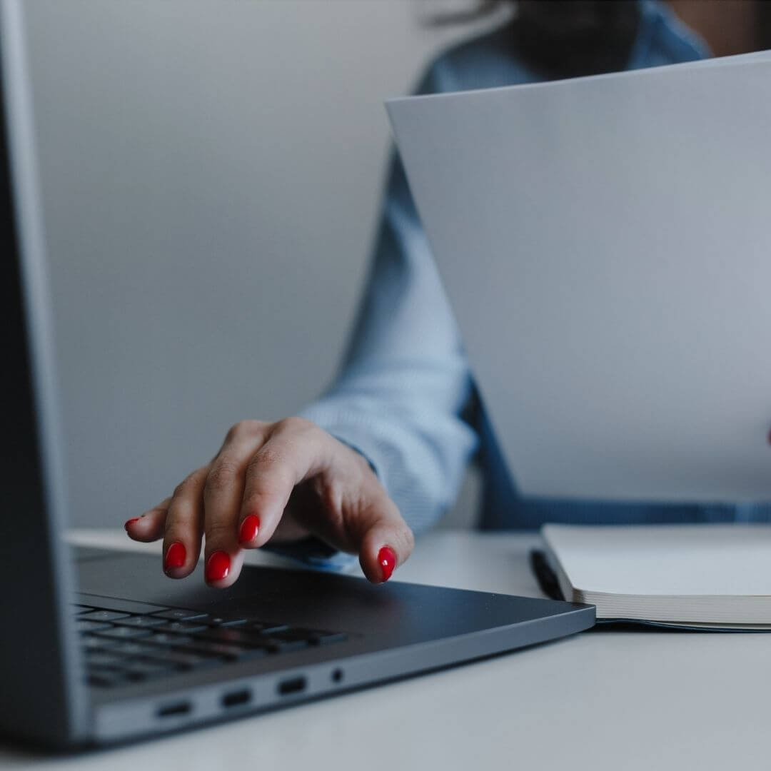 Mãos de uma profissional operando um notebook e manuseando papéis, simbolizando a organização técnica de documentos e ementas acadêmicas.
